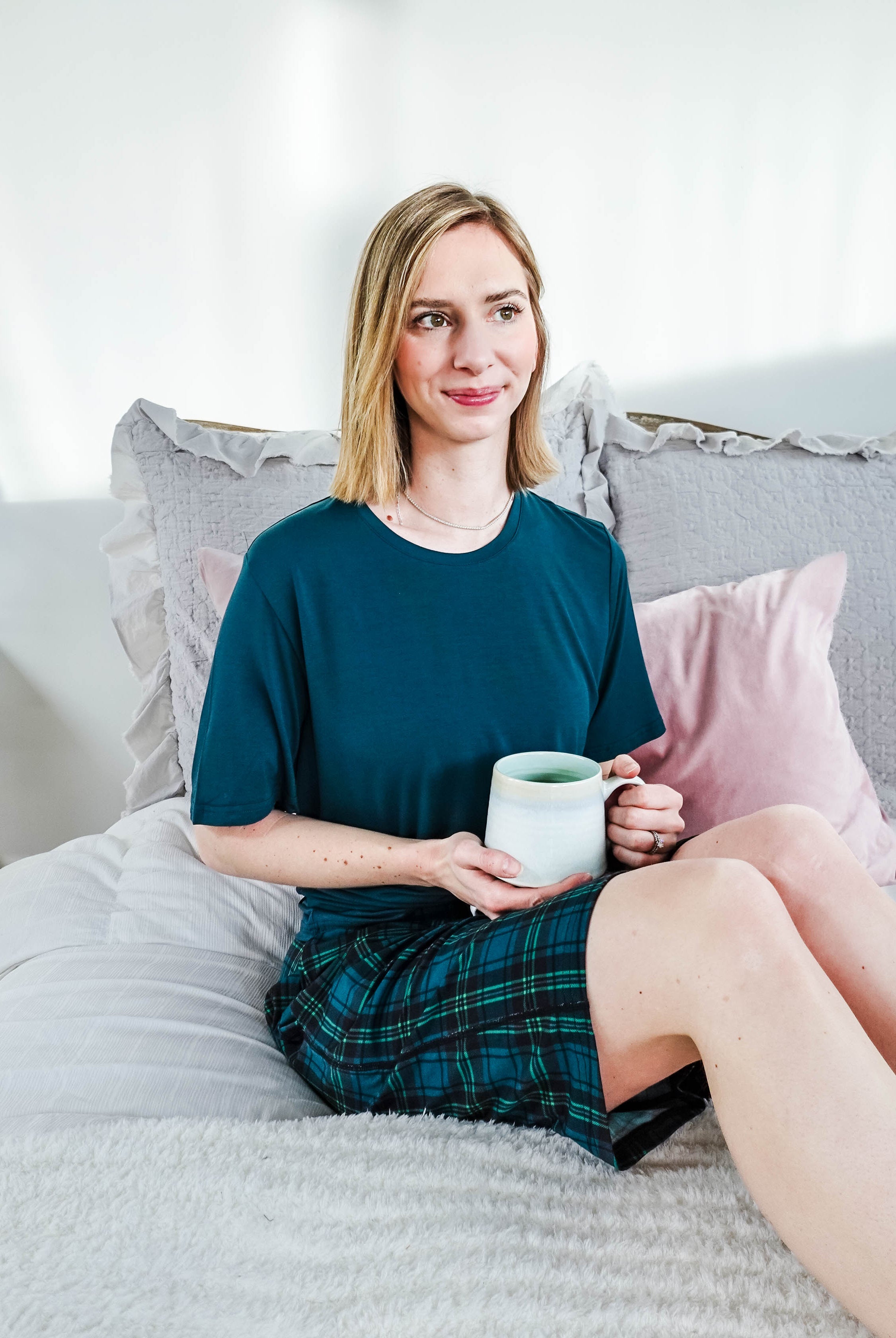 A woman sitting on a bed in Midnight Plaid Unisex Bamboo Sleep Shorts, holding a mug, enjoying a cozy atmosphere.