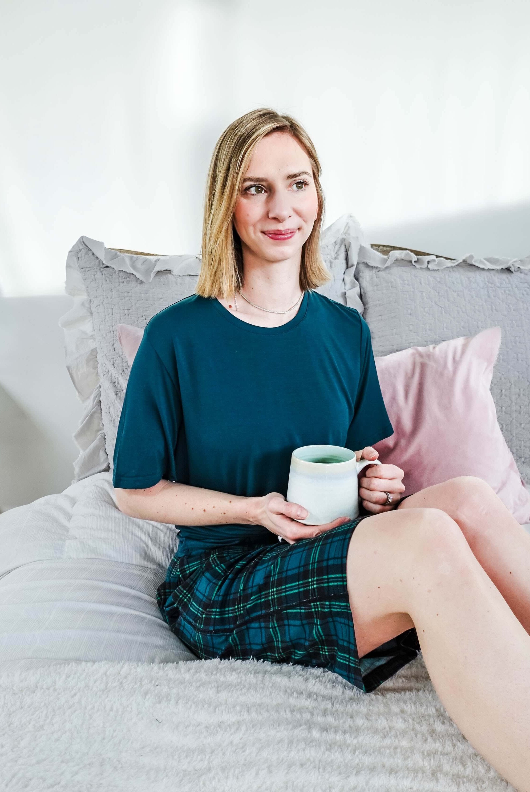 A woman sitting on a bed in Midnight Plaid Unisex Bamboo Sleep Shorts, holding a mug, enjoying a cozy atmosphere.