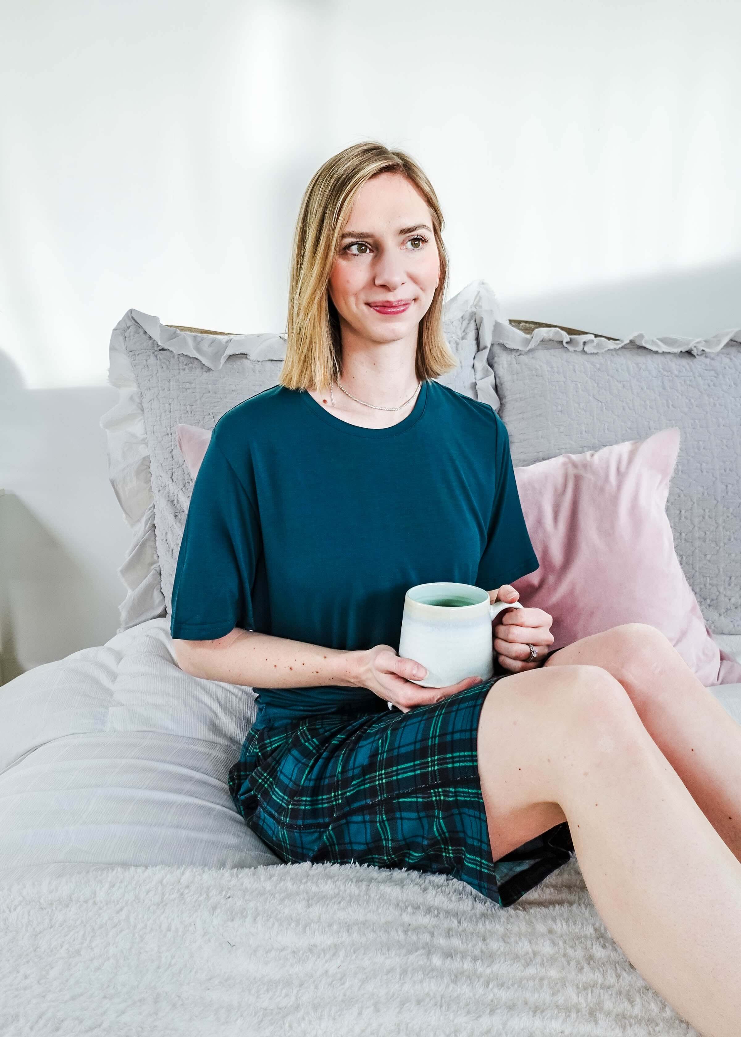 A woman sitting on a bed in Midnight Plaid Unisex Bamboo Sleep Shorts, holding a mug, enjoying a cozy atmosphere.