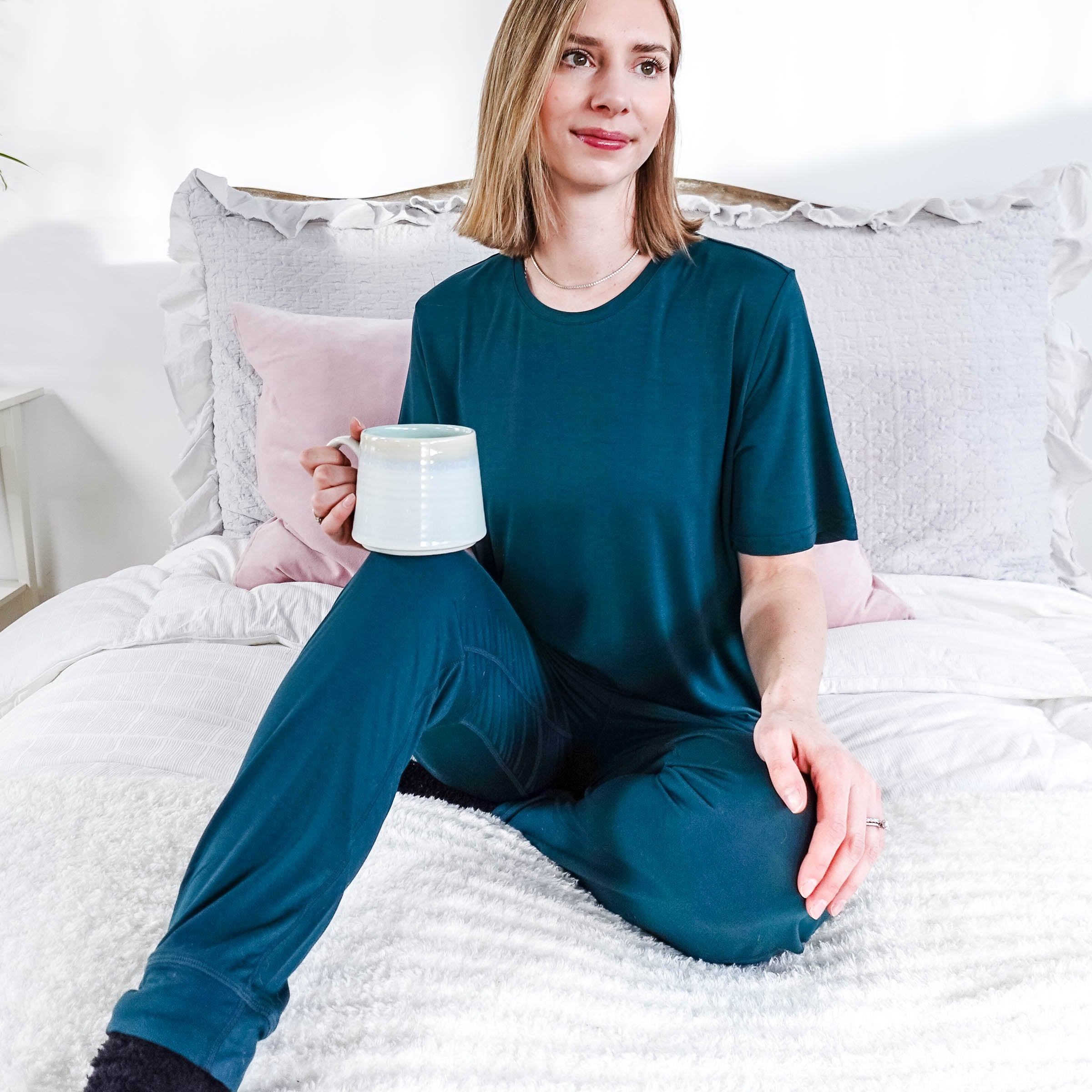 Model wearing Midnight Teal Unisex Bamboo Joggers while holding a cup and sitting on a bed.