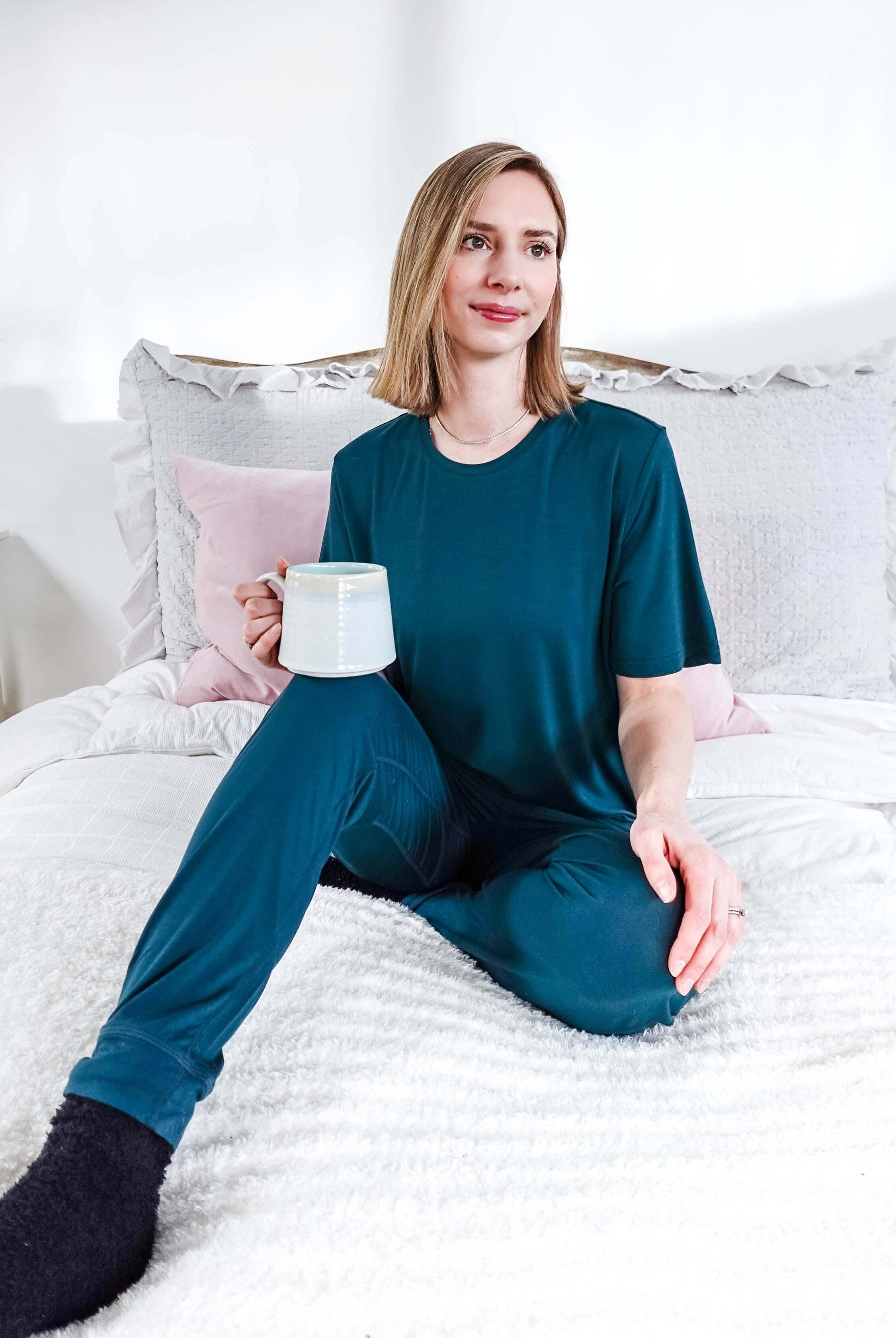 Model wearing Midnight Teal Unisex Bamboo Joggers while holding a cup and sitting on a bed.