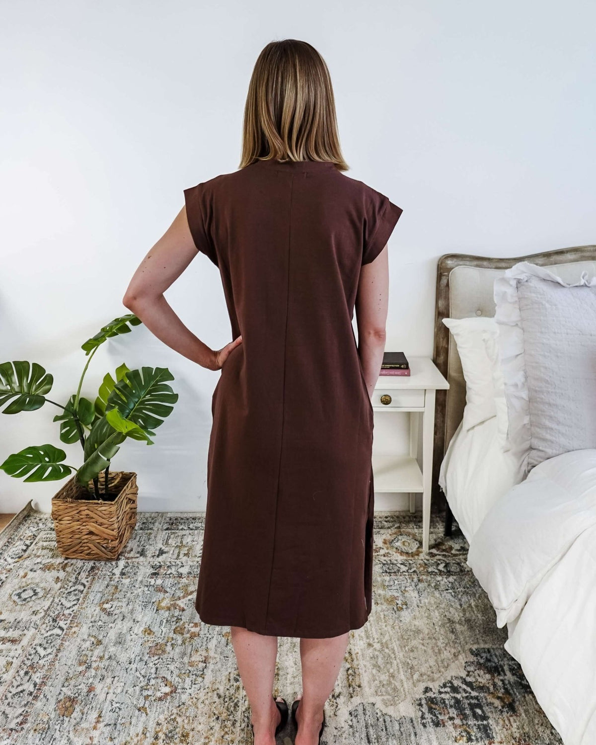 The back of a woman in a brown midi dress holding a beige handbag in a bedroom setting.