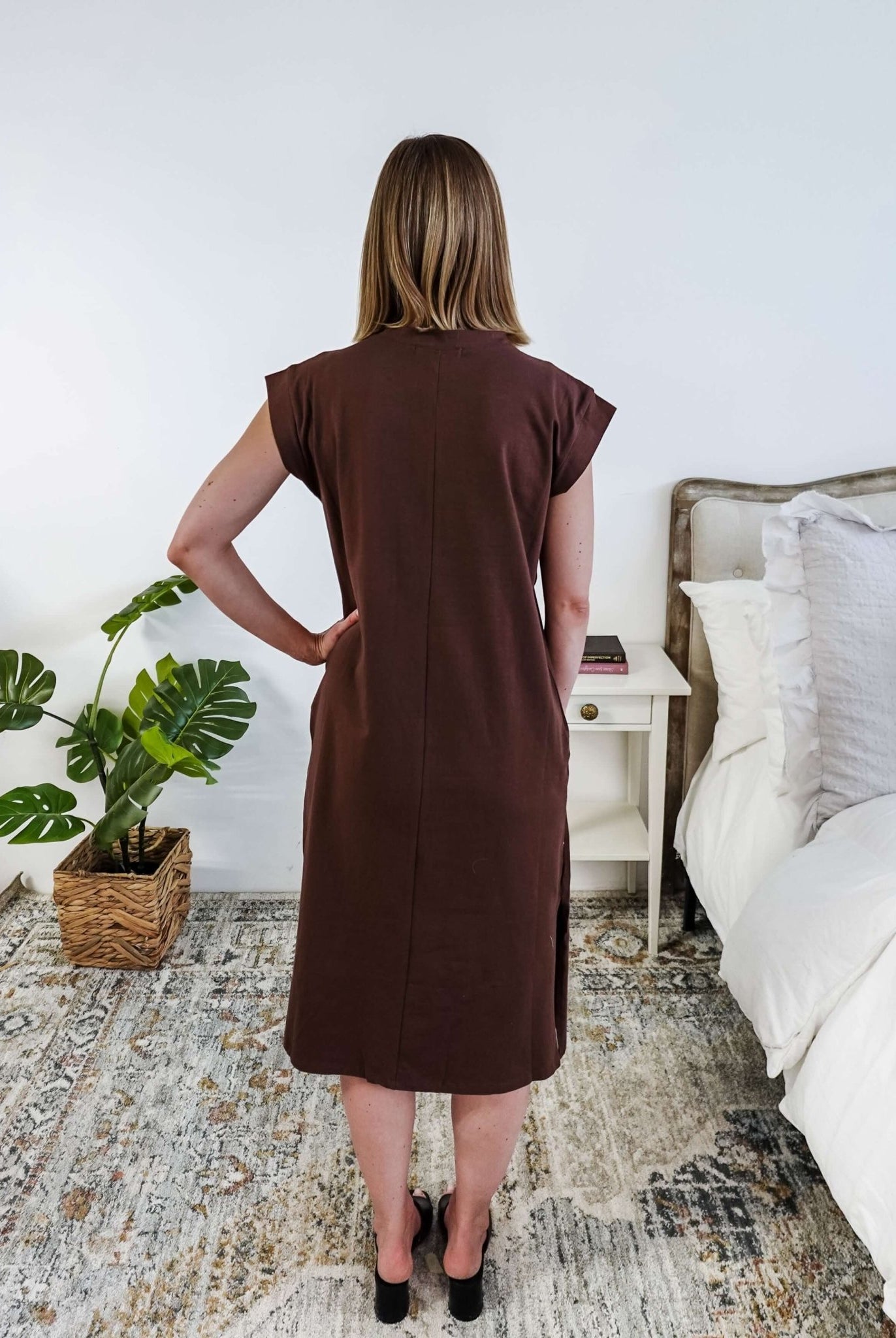 The back of a woman in a brown midi dress holding a beige handbag in a bedroom setting.