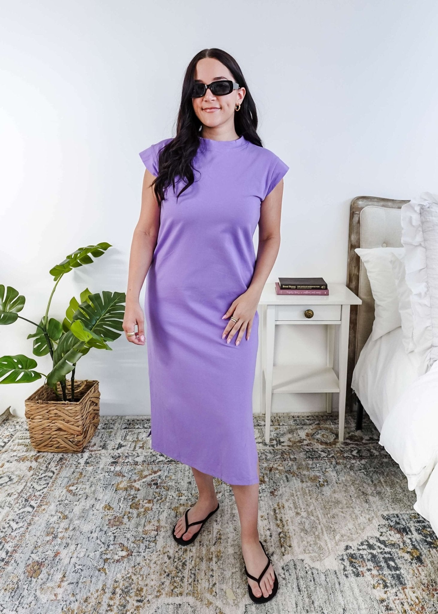 Woman in a purple midi dress holding a beige handbag in a bedroom setting.