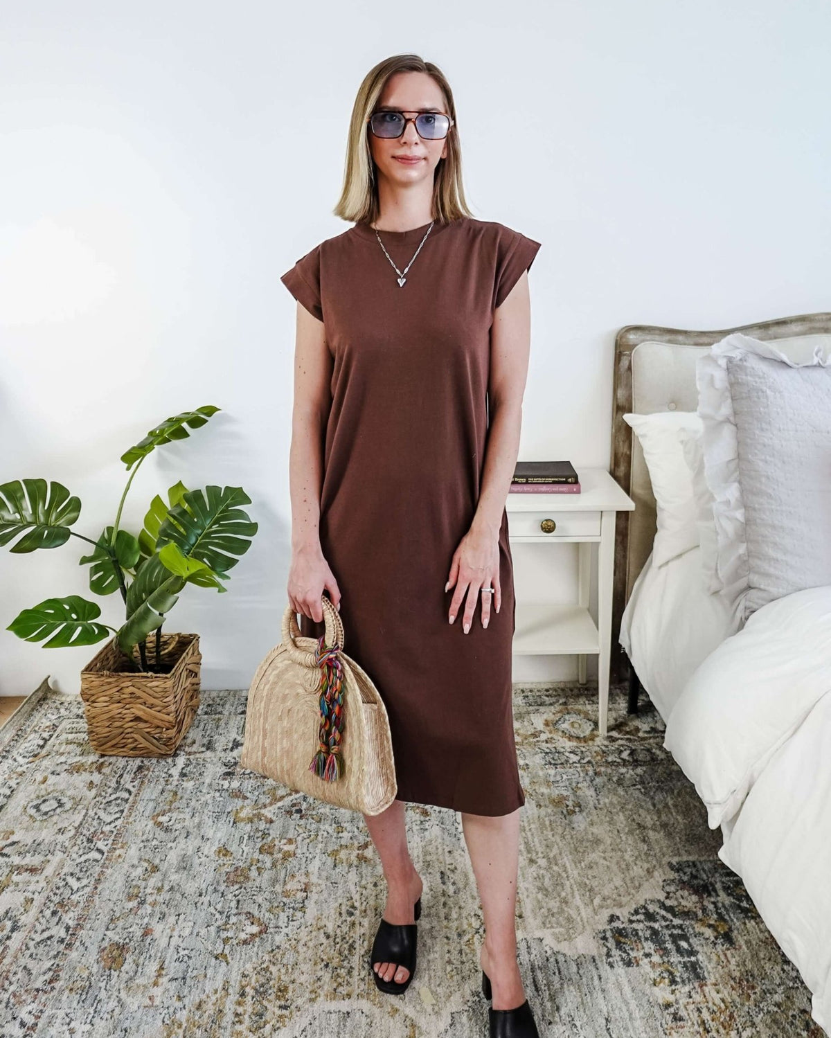Woman in a brown midi dress holding a beige handbag in a bedroom setting.