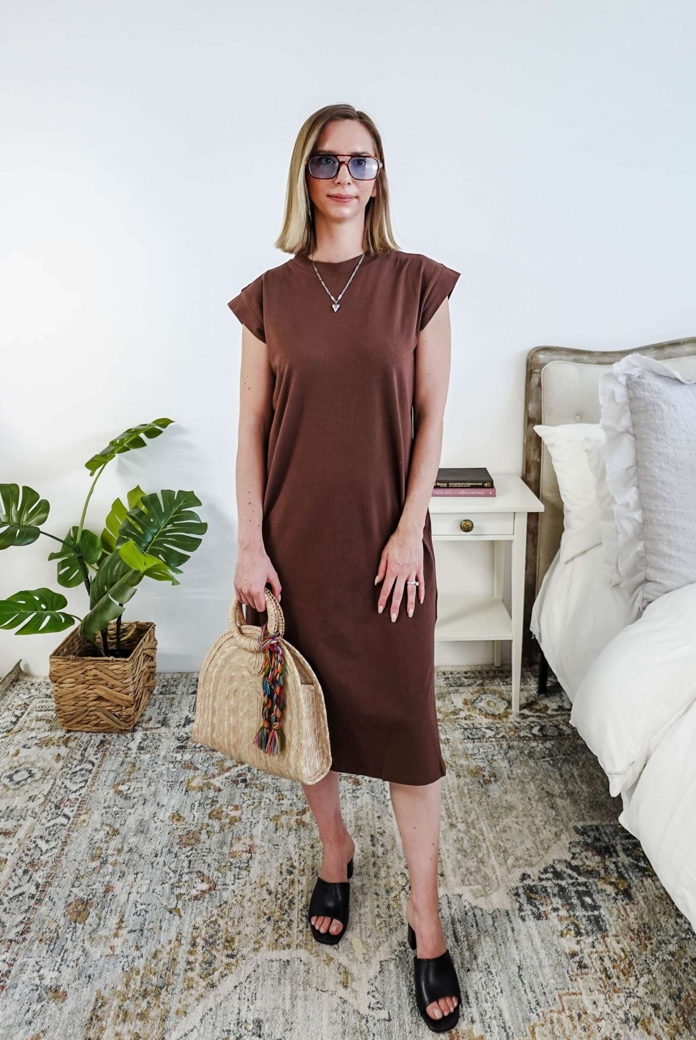 Woman in a brown midi dress holding a beige handbag in a bedroom setting.