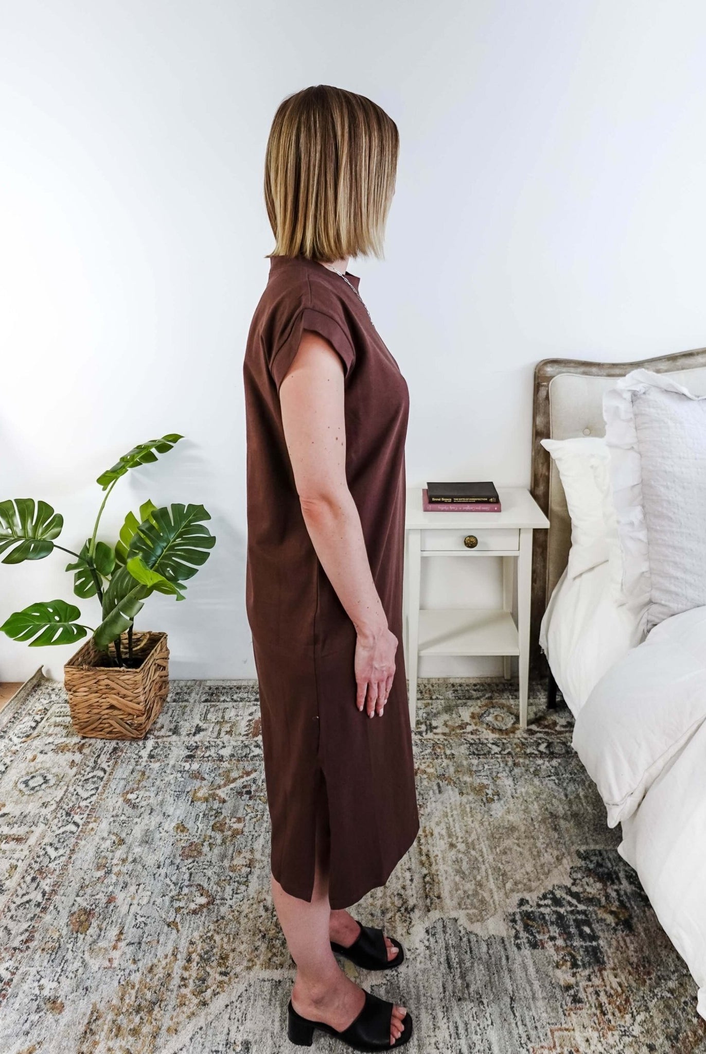 Woman in a brown midi dress holding a beige handbag in a bedroom setting.