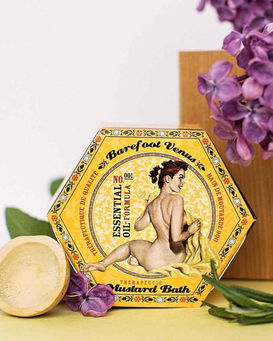 Mustard Bath Detox Bath Salts packaging featuring woman illustration, surrounded by purple flowers and a wooden spoon.