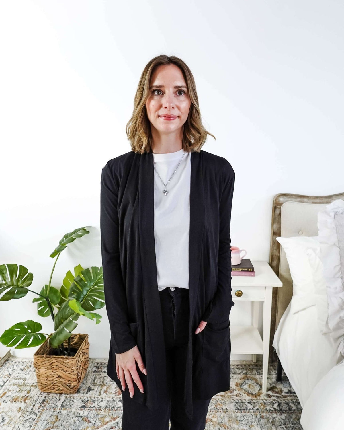 Woman in a black cardigan standing in a room with a plant and a bed in the background Scarlett Ellie Summer Open Lightweight Cardigan | Cloud Nine Pajamas