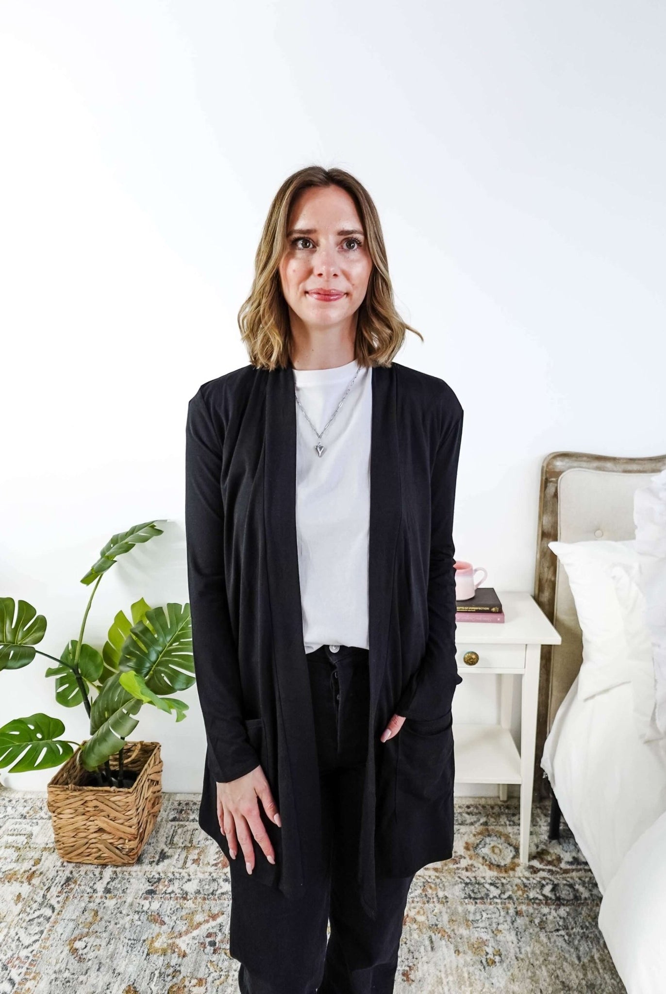 Woman in a black cardigan standing in a room with a plant and a bed in the background Scarlett Ellie Summer Open Lightweight Cardigan | Cloud Nine Pajamas