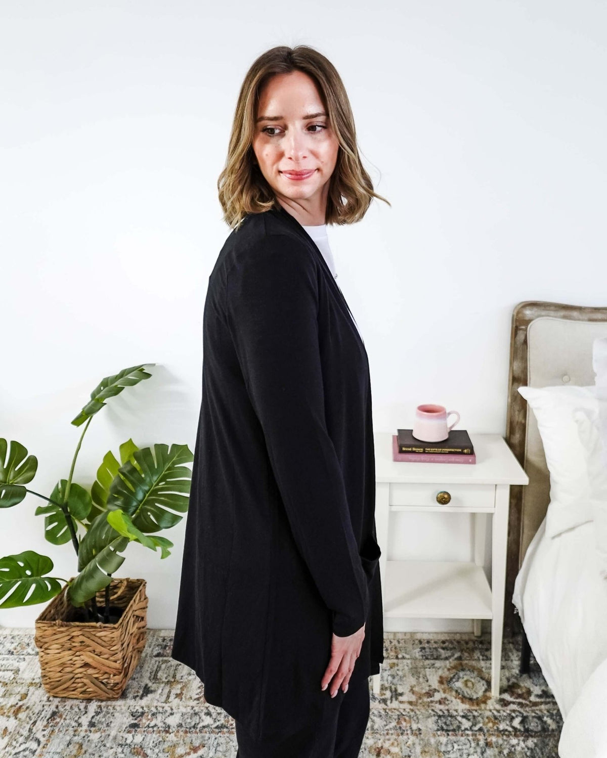 Woman in a black cardigan standing in a room with a plant and a bed in the background Scarlett Ellie Summer Open Lightweight Cardigan | Cloud Nine Pajamas