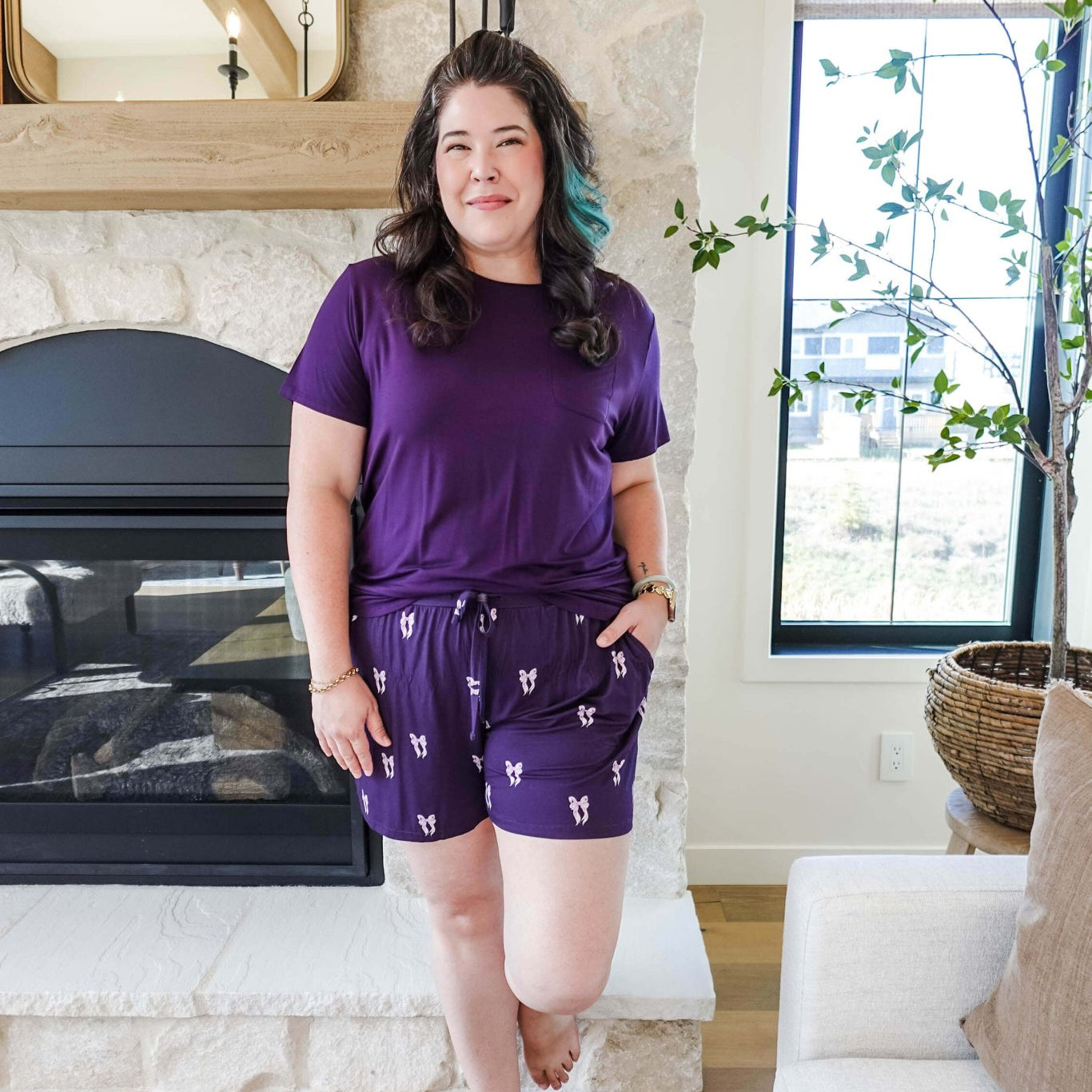 Woman wearing a Purple Bows Print Tee & Short Bamboo PJ Set by GEORGIA RYLAN, standing by a cozy fireplace.