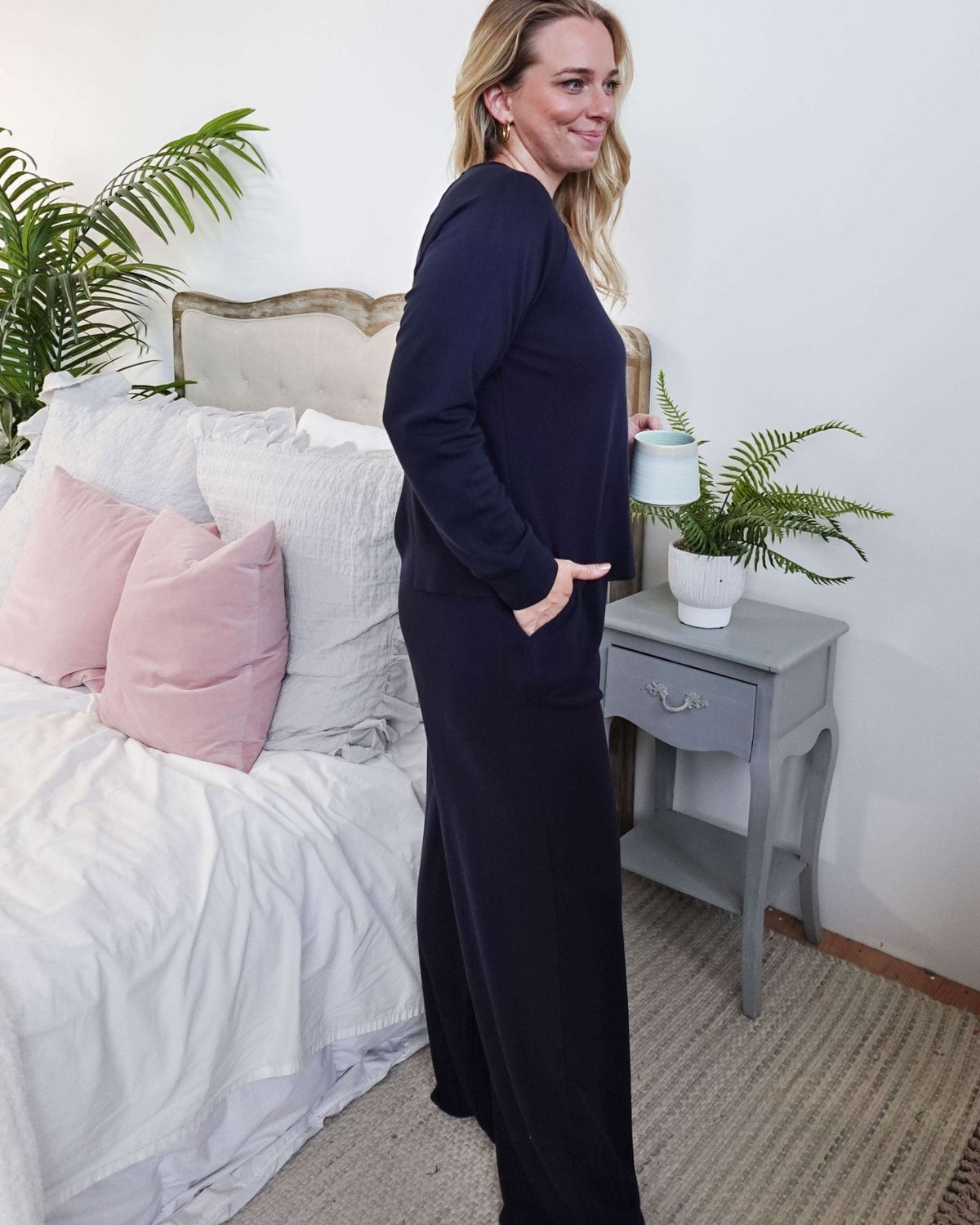 Woman in Long Sleeve Raw Edge Straight Pant Lounge Set standing by a bed, holding a drink, with a relaxed smile.