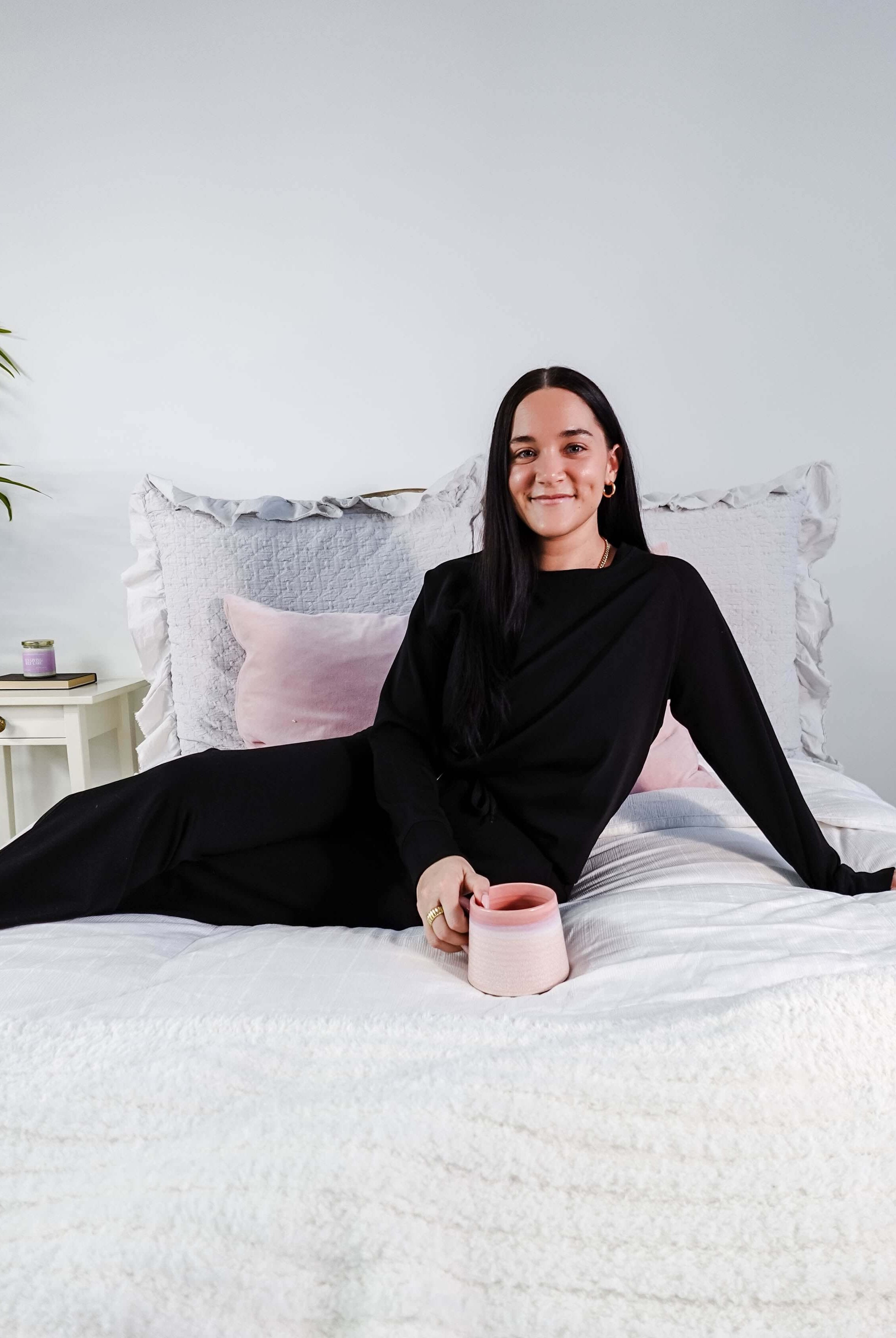 Model wearing Black Long Sleeve Raw Edge Straight Pant Cloud Lounge Set on a bed with a pink mug.