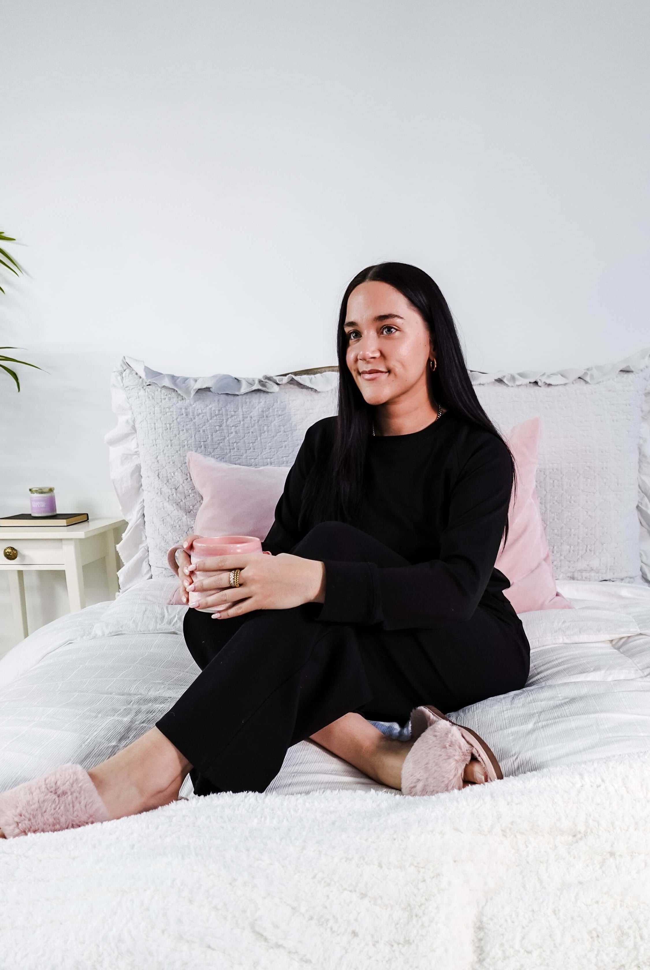 Woman wearing Black Long Sleeve Raw Edge Straight Pant Cloud Lounge Set seated on a bed holding a drink.