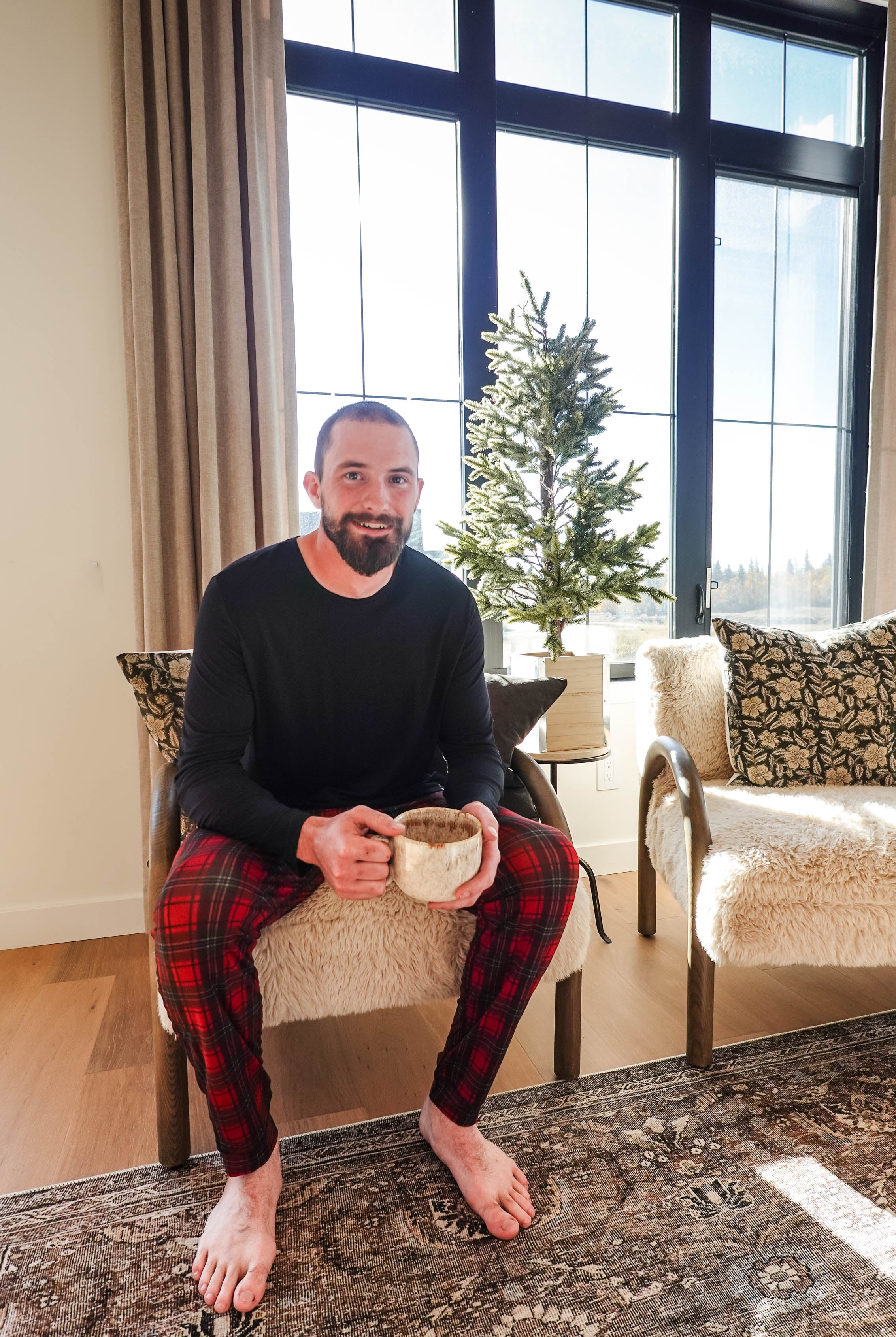 A man wearing Red Plaid Unisex Bamboo Jogger from Bob's Sleepwear Co. sitting with a mug in cozy interior.