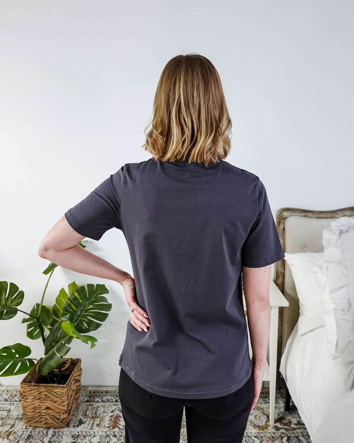 A lady standing in a bedroom in a dark grey shirt.