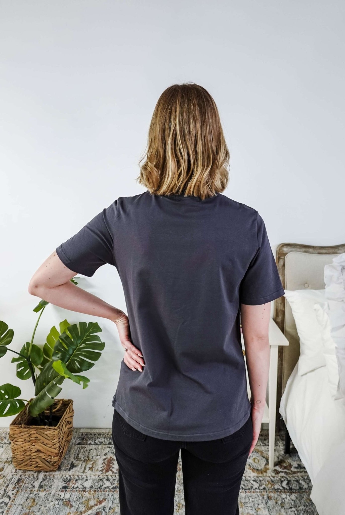 A lady standing in a bedroom in a dark grey shirt.
