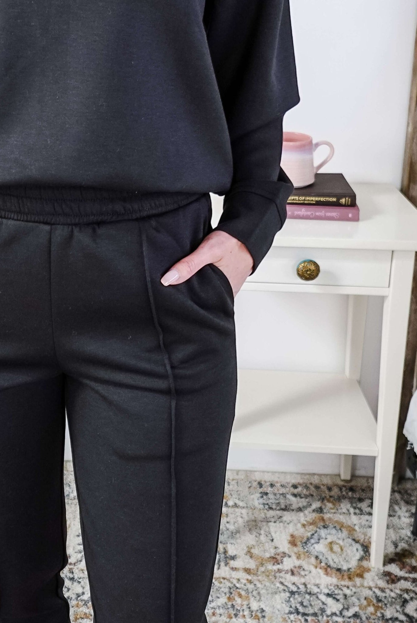 Person wearing black pants with a drawstring waistband, blurred background of a white shelf and books.