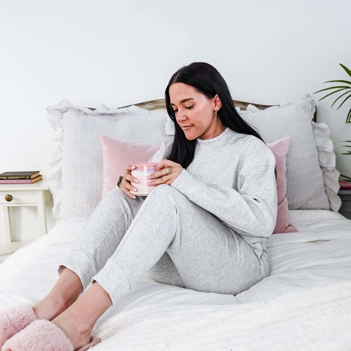Woman in gray pajamas sitting on a bed holding a pink cup, surrounded by pillows and a plant.