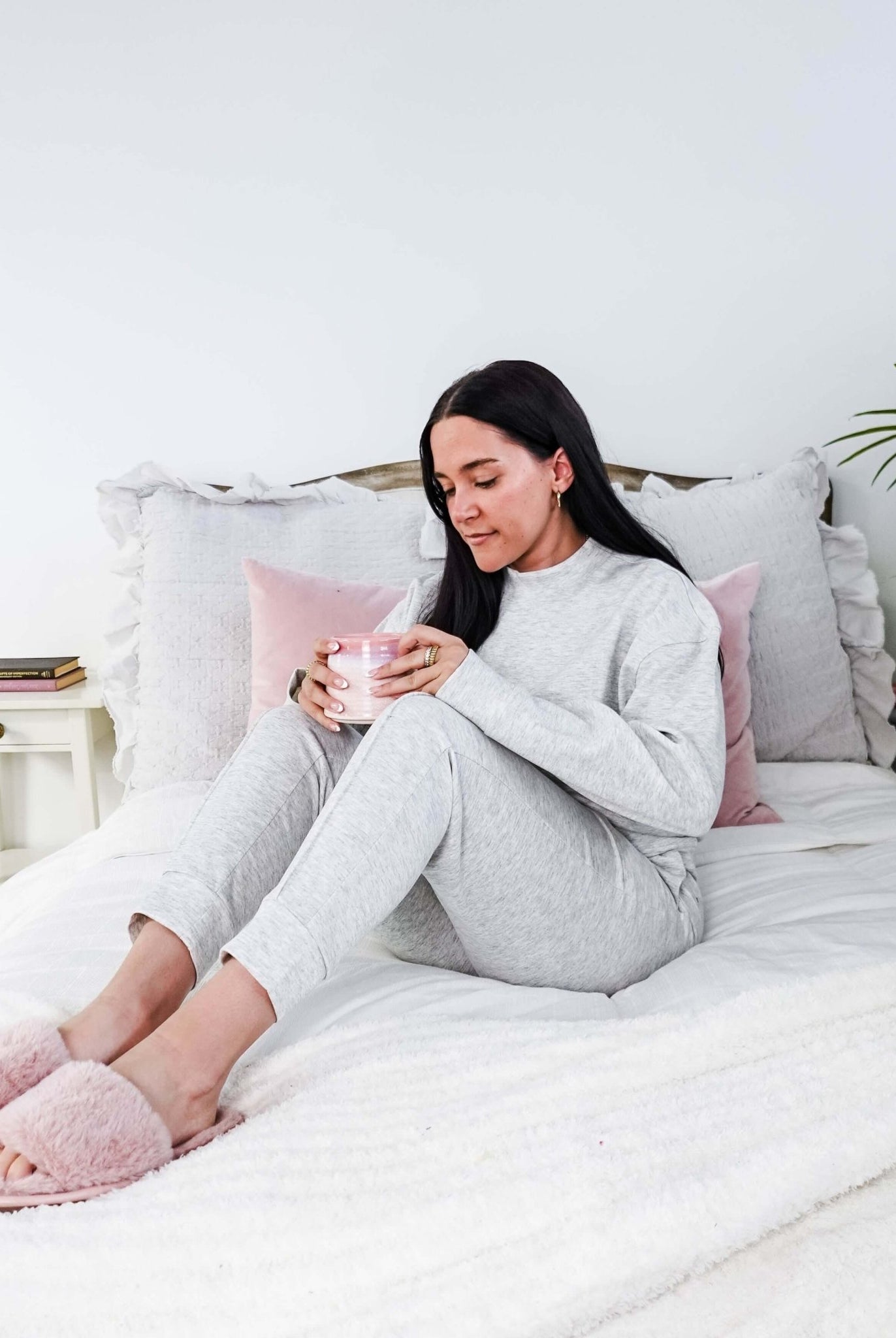 Woman in gray pajamas sitting on a bed holding a pink cup, surrounded by pillows and a plant.