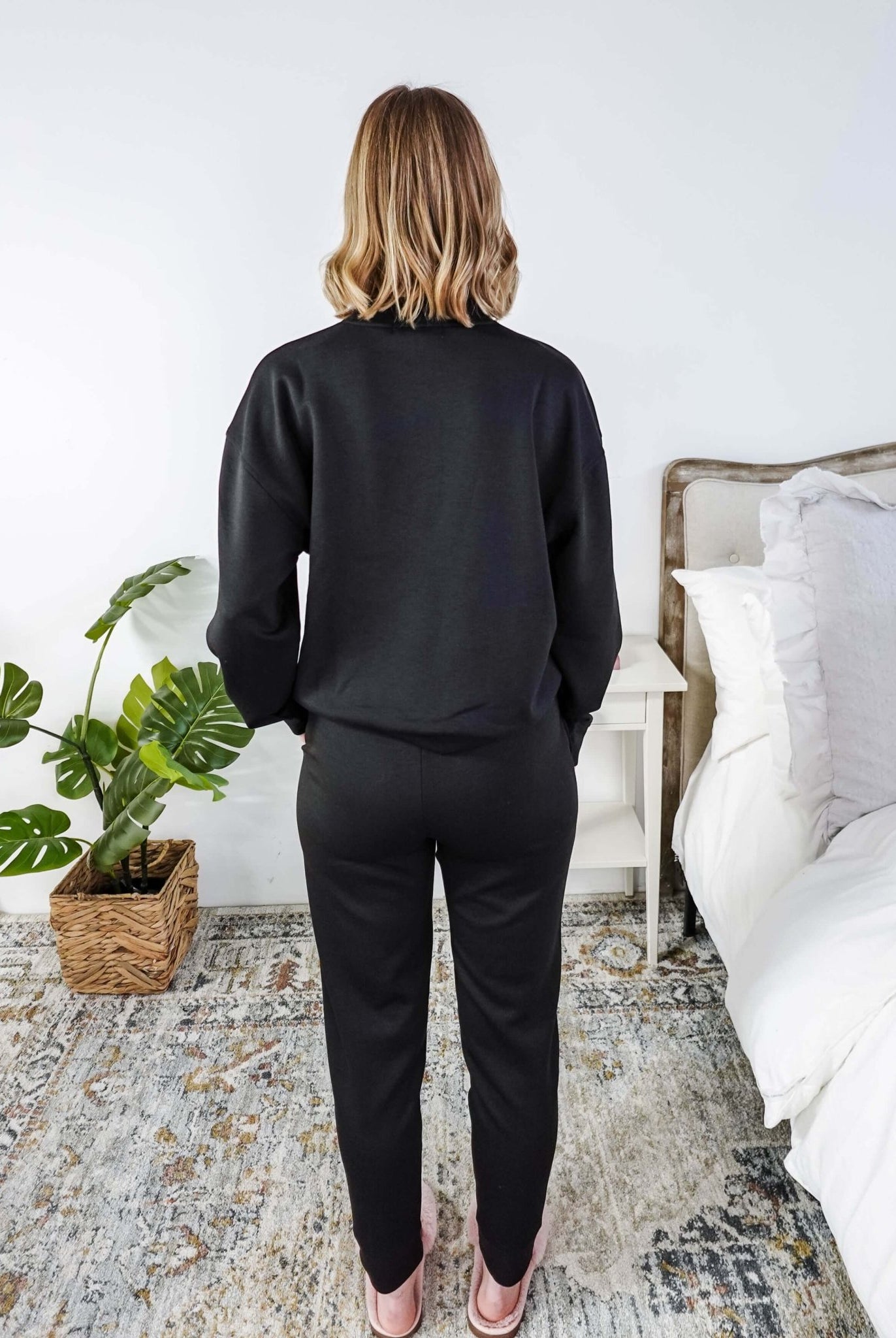 Person wearing a black outfit standing in a bedroom with a plant and bed in the background.
