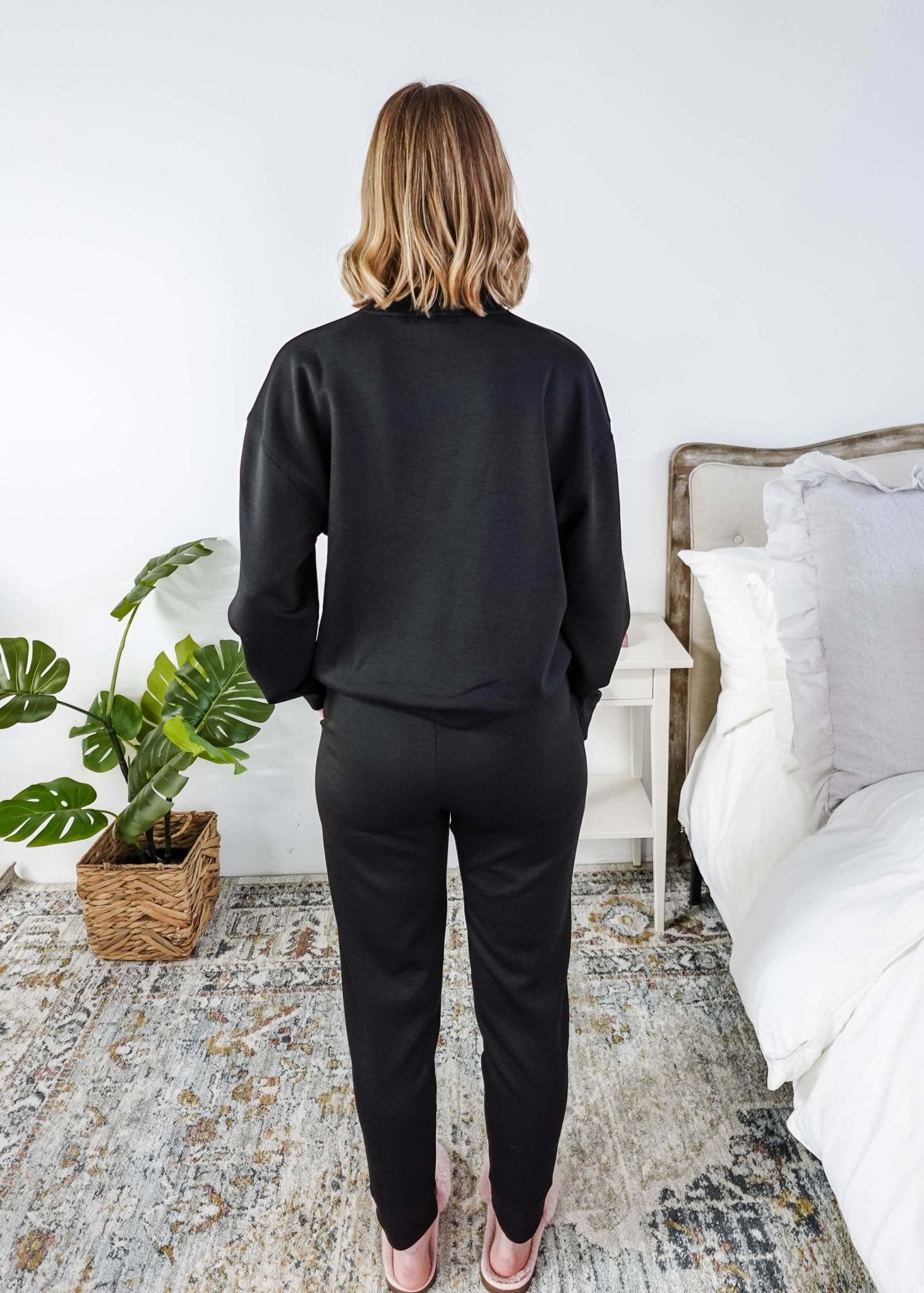 Person wearing a black outfit standing in a bedroom with a plant and bed in the background.