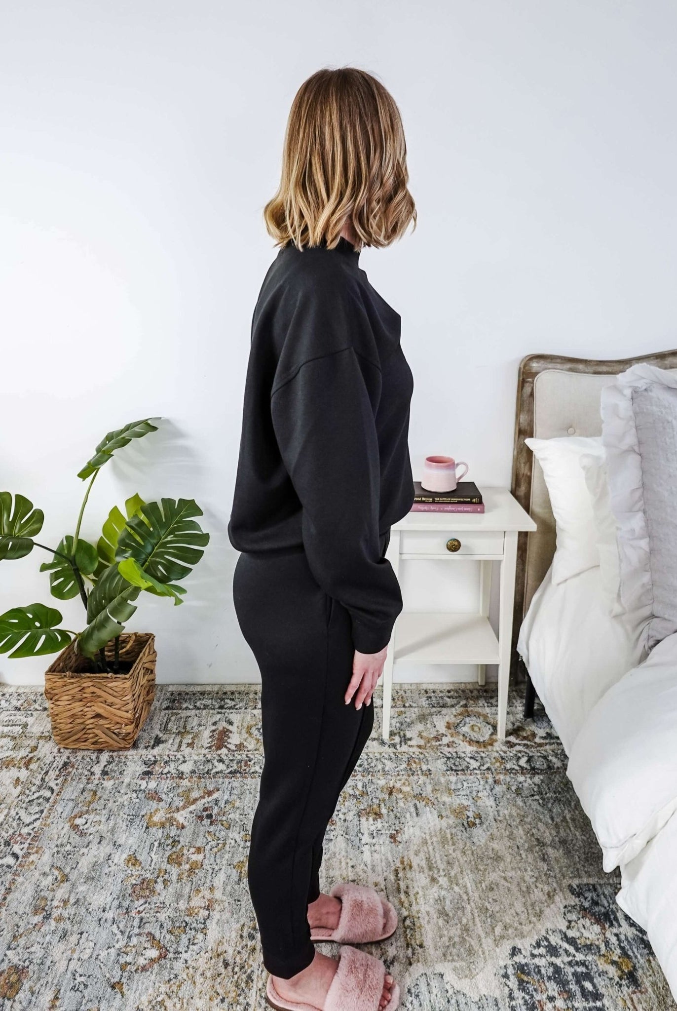 Woman in black tracksuit standing in a bedroom with a plant and bed in the background