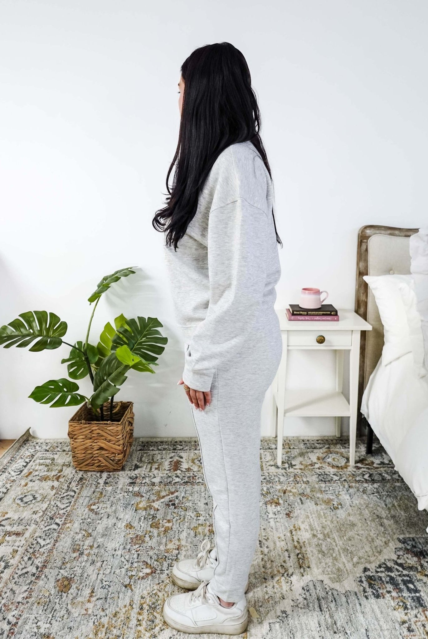 Woman in a cozy room with a plant and a nightstand wearing a grey lounge set.