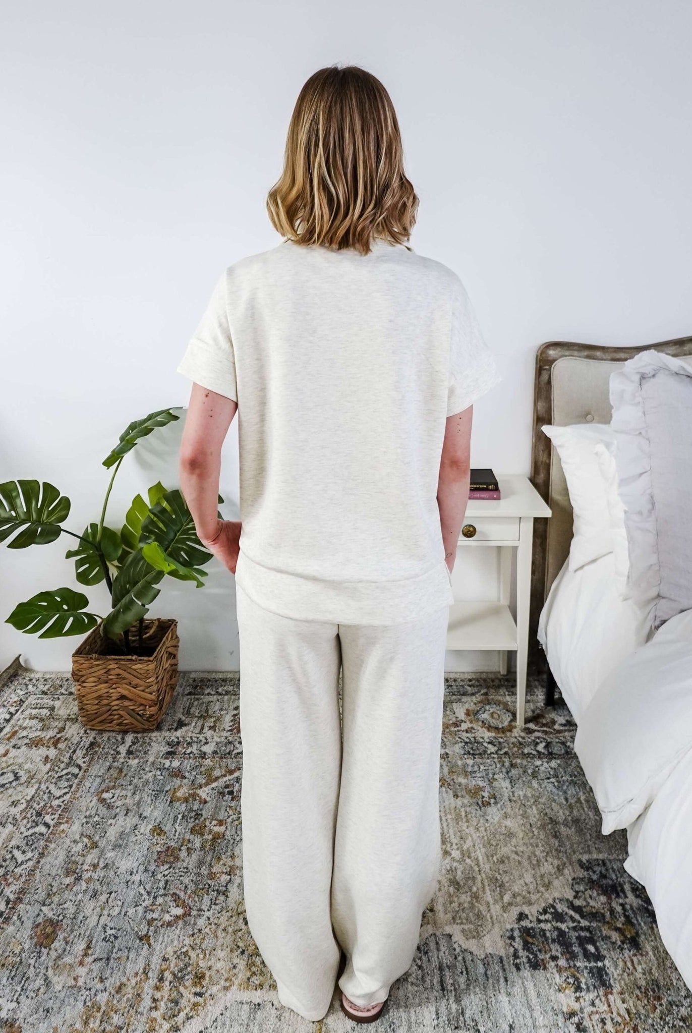 The back of a woman in a cream lounge outfit standing in a bedroom with a plant and bed in the background