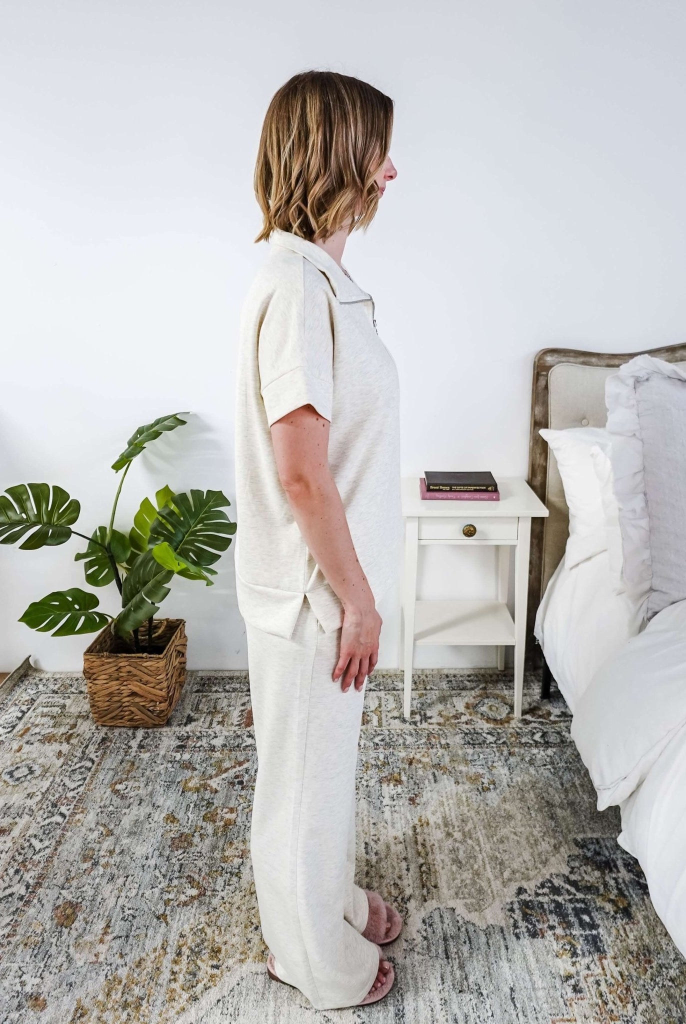 Woman in a cream lounge outfit standing in a bedroom with a plant and bed in the background