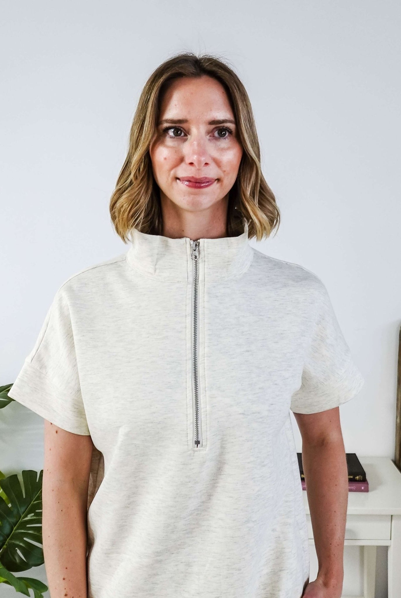 Woman in a cream lounge outfit standing in a bedroom with a plant and bed in the background. Sweater is zipped all the way up.