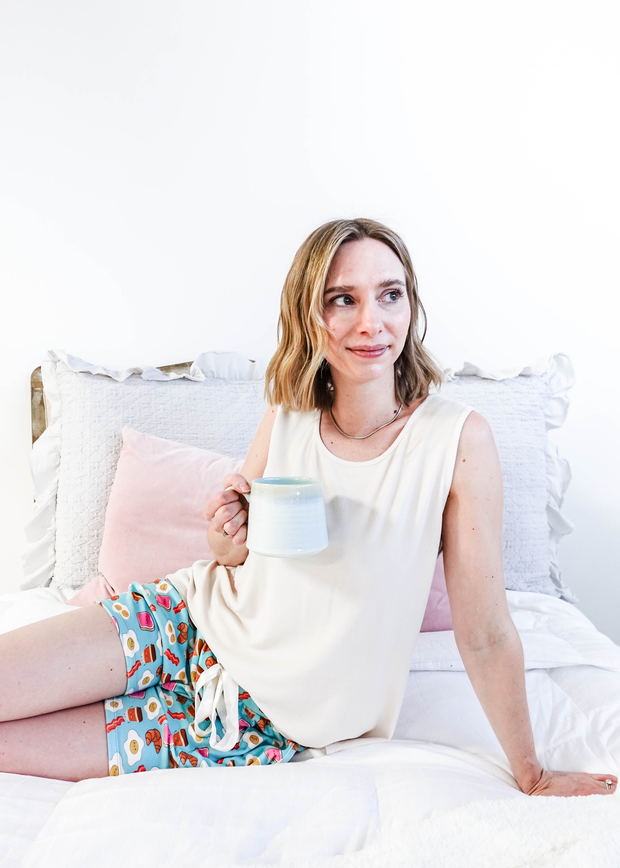 Lifestyle shot of Miranda on a bed wearing the Brunch Club velour shorts with eggs and bacon print and ivory peachy tank.
