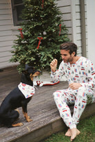A man in festive pajamas interacts with a dog wearing a Holiday Print Bamboo Pet Bandana near a Christmas tree.