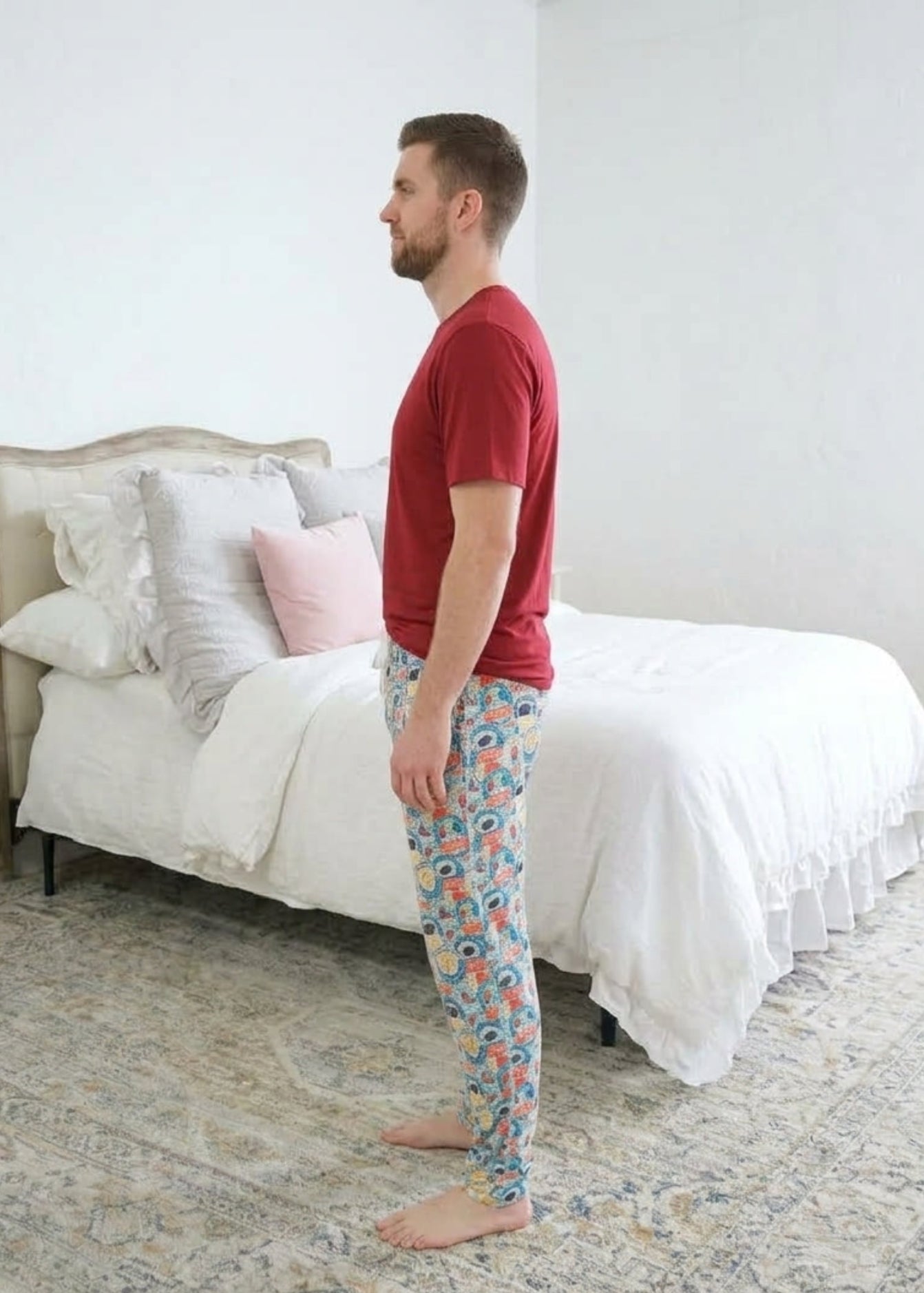 A man standing sideways wearing a red bamboo short sleeve t-shirt and colorful hockey patterned pajama pants stands barefoot in a bedroom with a white bed and patterned rug. Cloud Nine Pajamas 