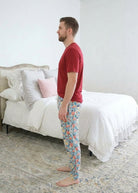 A man standing sideways wearing a red bamboo short sleeve t-shirt and colorful hockey patterned pajama pants stands barefoot in a bedroom with a white bed and patterned rug. Cloud Nine Pajamas 