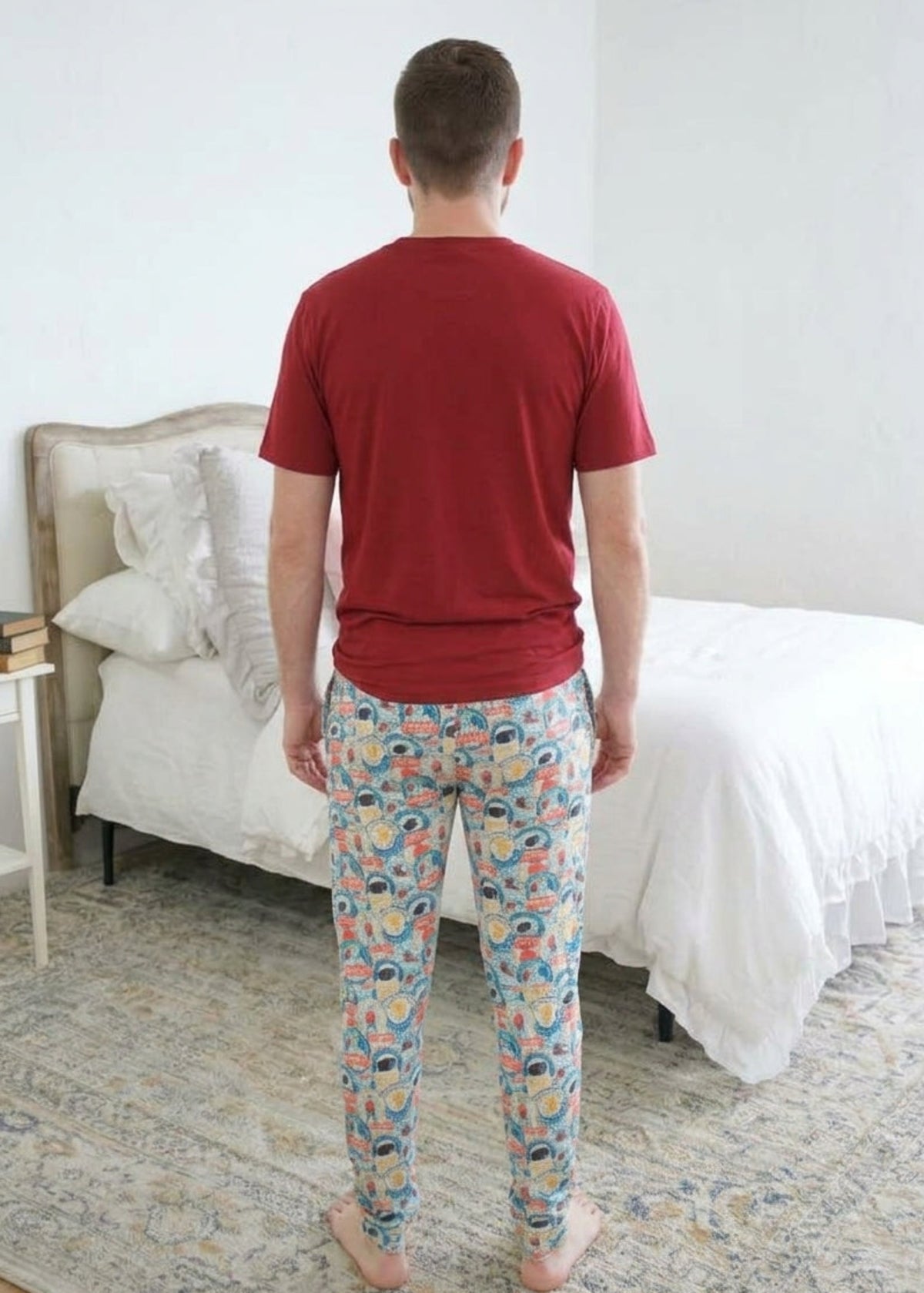 A man standing backwards wearing a red bamboo short sleeve t-shirt and colorful hockey patterned pajama pants stands barefoot in a bedroom with a white bed and patterned rug. Cloud Nine Pajamas 