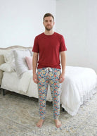 A man wearing a red bamboo short sleeve t-shirt and colorful hockey patterned pajama pants stands barefoot in a bedroom with a white bed and patterned rug. Cloud Nine Pajamas Edmonton