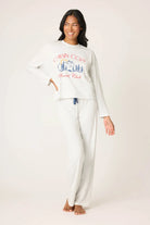 Woman wearing a white long-sleeve top and pants with a cabin cozy social club graphic design on a plain background