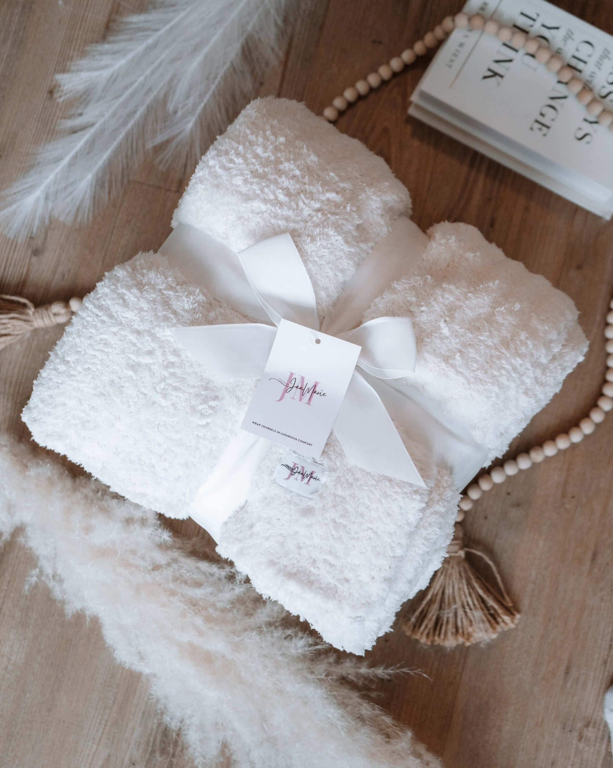 Cozy Cabin Blanket – Oversized Ivory Plush Throw (75″ × 52″). Plush ivory white blanket shown folded in a square and wrapped with a satin ribbon in a lay flat setting. There are wooden beads, books and pampas grass shown around the blanket aesthetically. The label and tag are shown. 