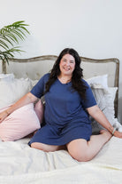 Stylish model wearing Denim Blue V Neck Bamboo Nightshirt dress with pockets, showcasing versatility in sleepwear and everyday fashion. Justine Mah lounges in bed wearing the dress.