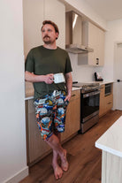 Man standing in kitchen wearing Mountains Are Calling Print Unisex Shorts Bamboo and a green t-shirt, holding a coffee cup.