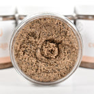 Coffee Bean Sugar Scrub in a clear jar showing the textured scrub, surrounded by blurred containers.