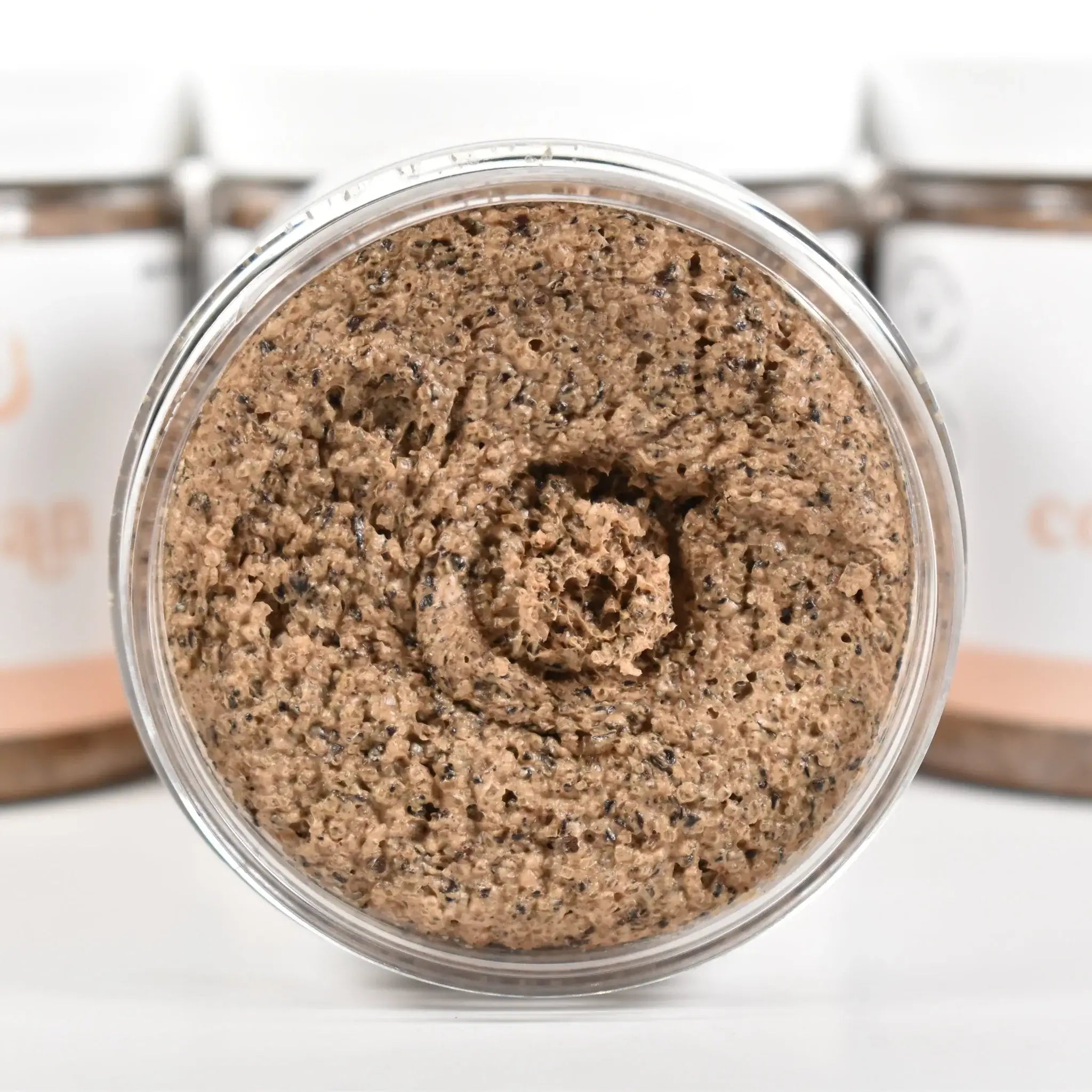 Coffee Bean Sugar Scrub in a clear jar showing the textured scrub, surrounded by blurred containers.
