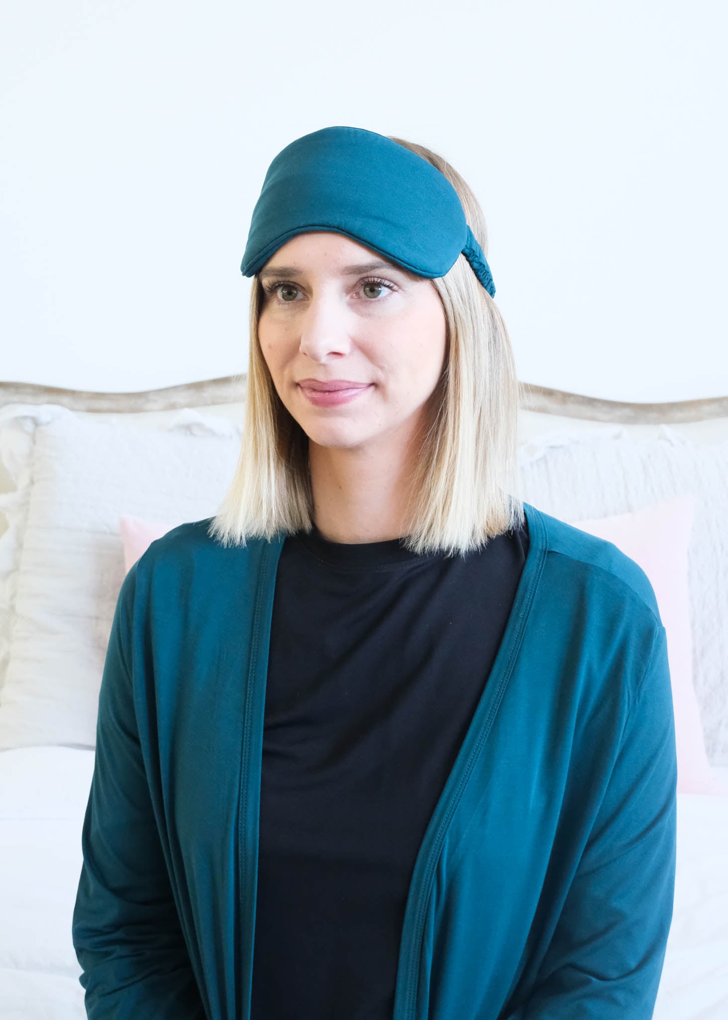 A woman with blonde shoulder-length hair wearing a dark teal sleep mask and a matching dark teal cardigan over a black shirt, posed in front of a light-colored bed.