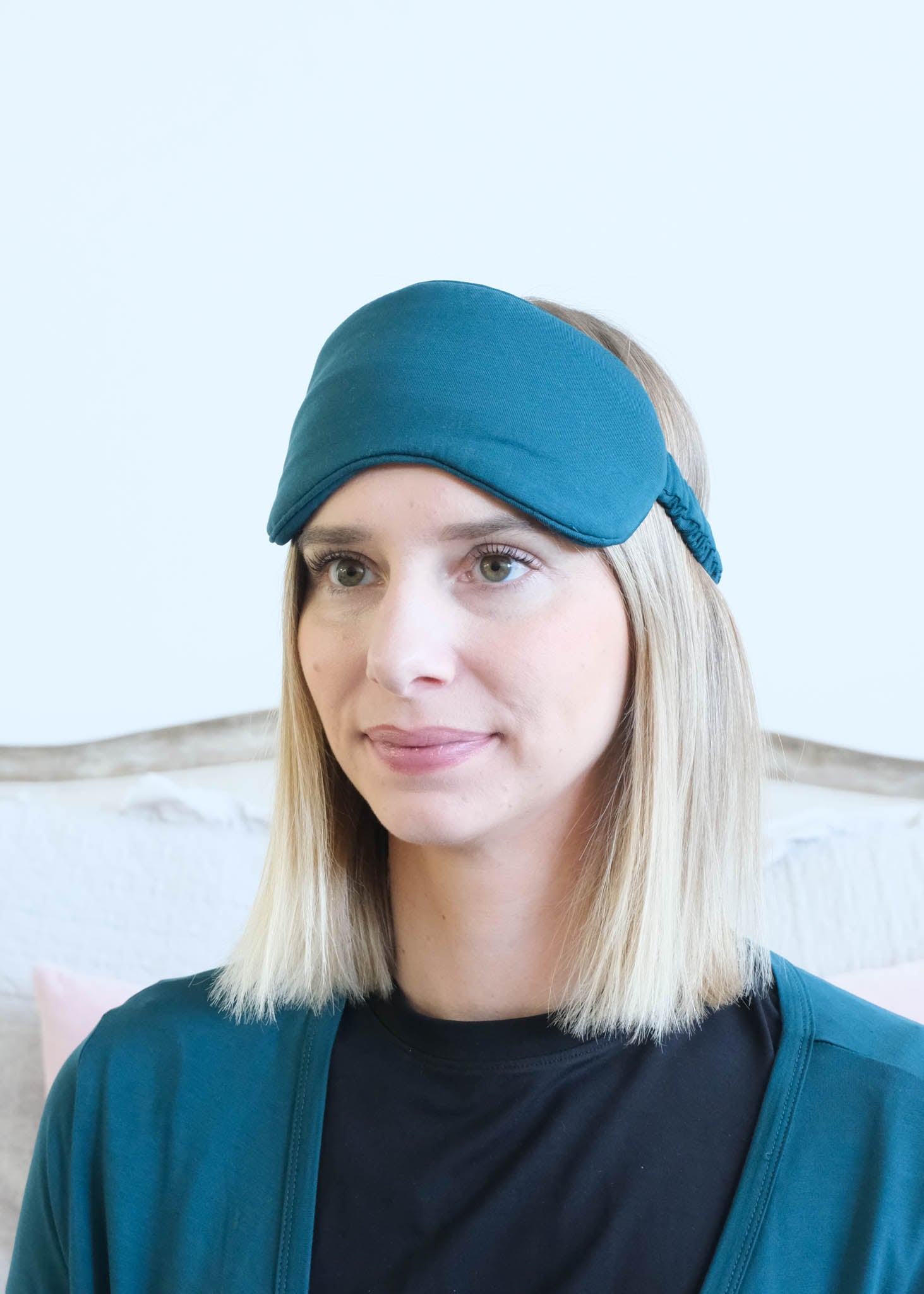 A woman with blonde hair wearing a dark teal sleep mask and a matching dark teal cardigan over a black shirt, set against a light-colored wall and a wooden headboard.