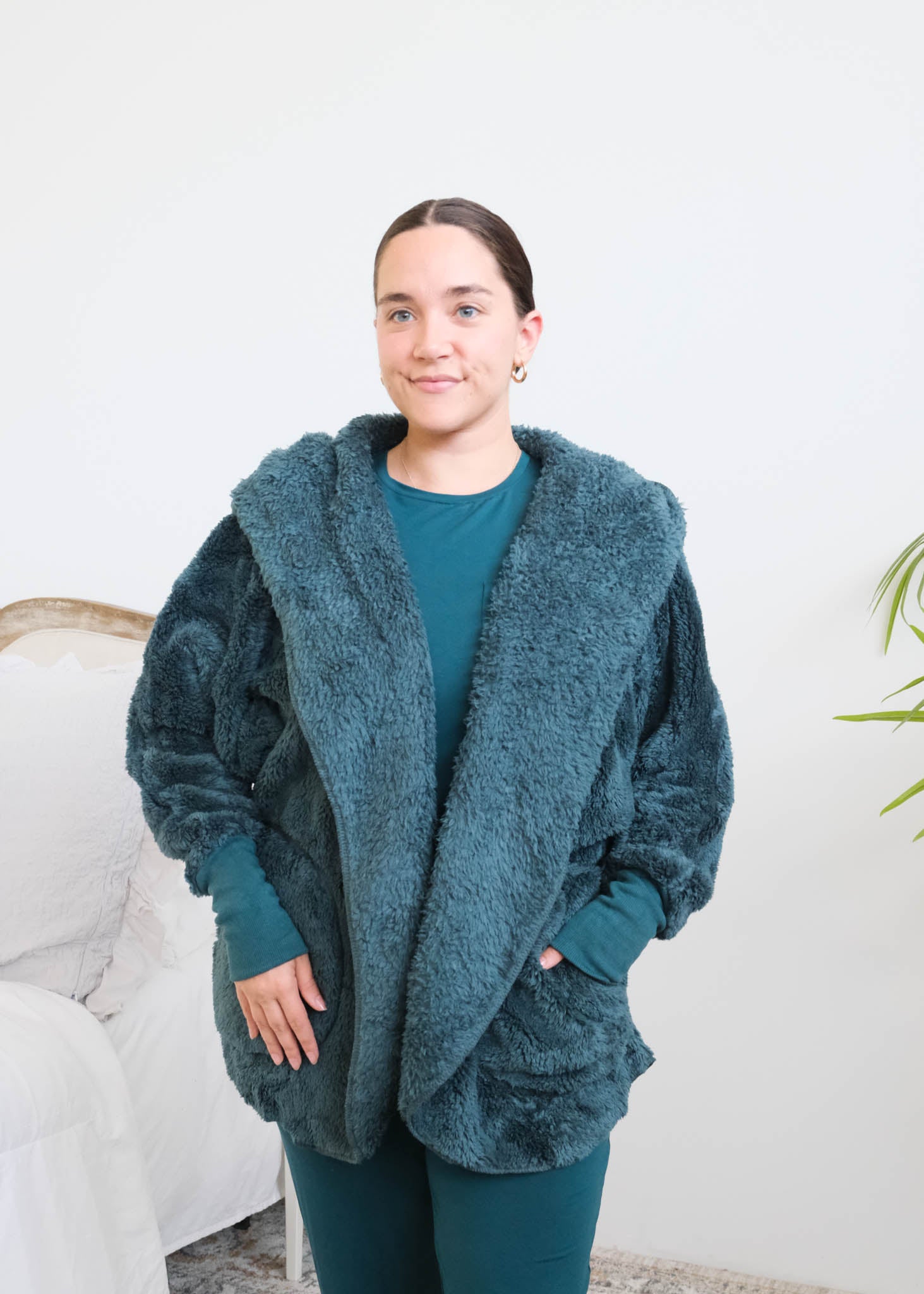 Close up of woman standing in a bedroom wearing a dark peacock teal fluffy hooded cardigan by Lounge by Jan Marie over a dark midnight teal bamboo pajamas, with ribbed cuffs and pink fuzzy slippers, hands tucked into the cardigan's front pockets. Cloud Nine Pajamas Edmonton