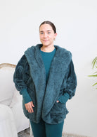 Close up of woman standing in a bedroom wearing a dark peacock teal fluffy hooded cardigan by Lounge by Jan Marie over a dark midnight teal bamboo pajamas, with ribbed cuffs and pink fuzzy slippers, hands tucked into the cardigan's front pockets. Cloud Nine Pajamas Edmonton