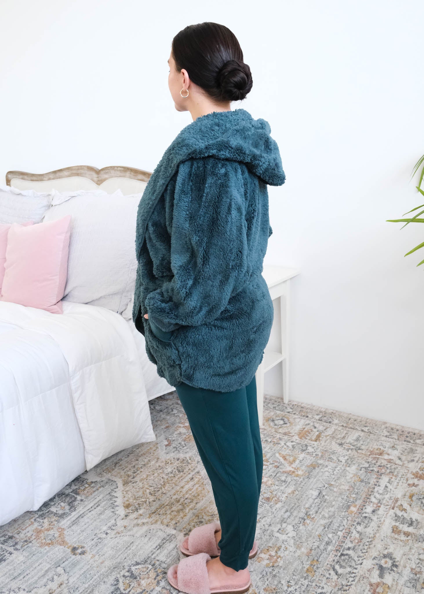 Women standing sideways in a bedroom wearing a dark peacock teal fluffy hooded cardigan by Lounge by Jan Marie over a dark midnight teal bamboo pajamas, with ribbed cuffs and pink fuzzy slippers, hands tucked into the cardigan's front pockets. Cloud Nine Pajamas Edmonton