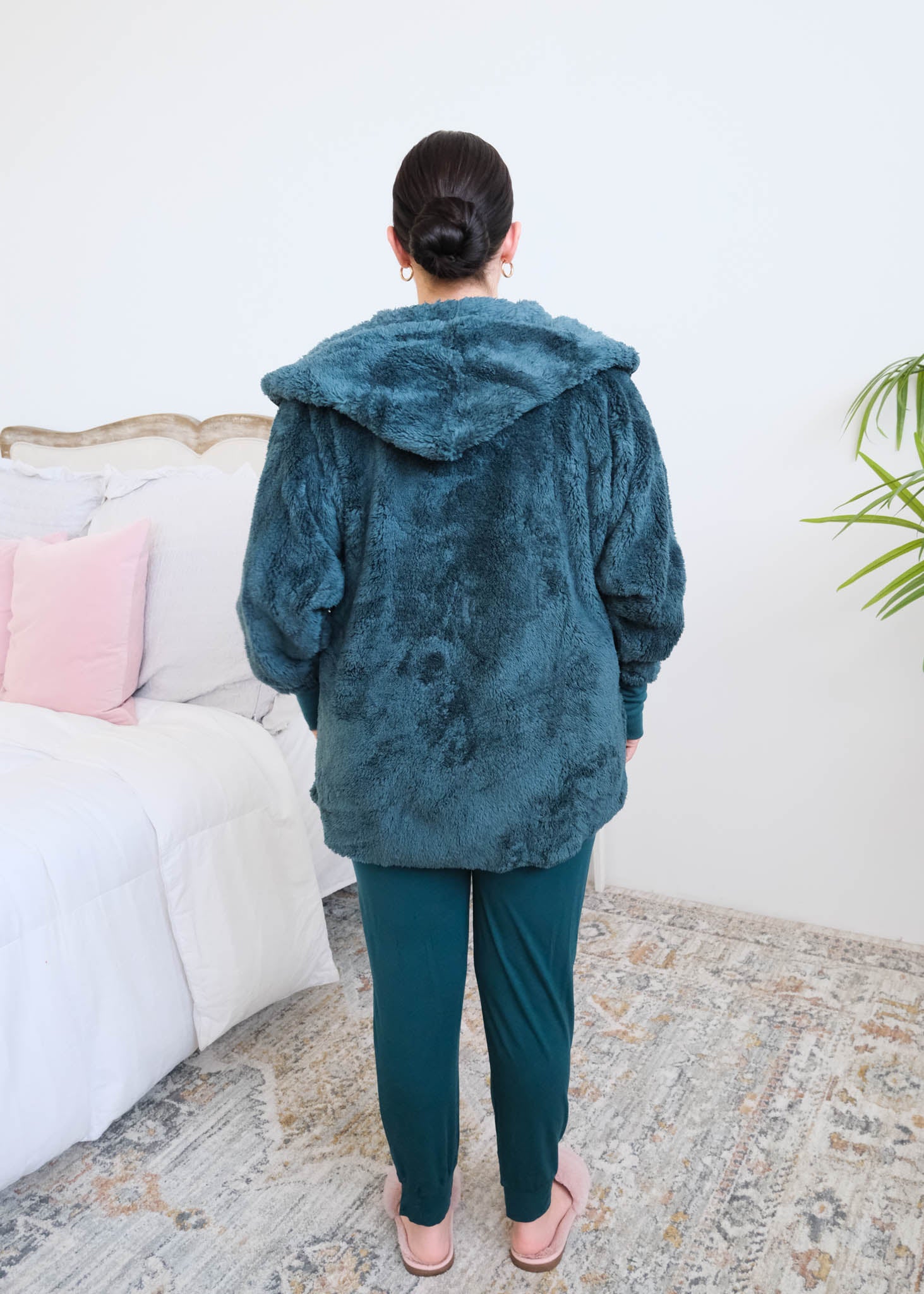 Woman standing backwards in a bedroom wearing a dark peacock teal fluffy hooded cardigan by Lounge by Jan Marie over a dark midnight teal bamboo pajamas, with ribbed cuffs and pink fuzzy slippers, hands tucked into the cardigan's front pockets. Cloud Nine Pajamas Edmonton